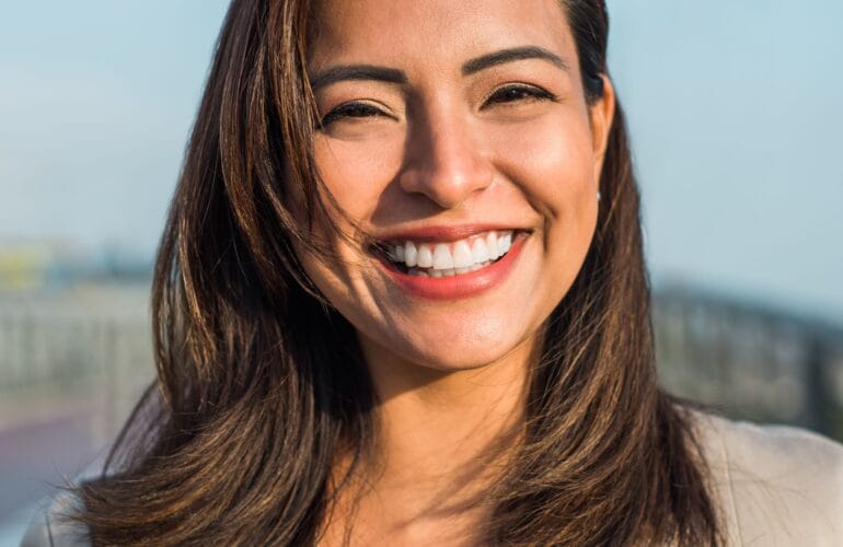 Mujer sonriente mostrando una sonrisa saludable y natural, con el cabello castaño al viento en un entorno exterior luminoso, representando el resultado ideal del tratamiento con los dentistas en Valladolid de la clínica MYM